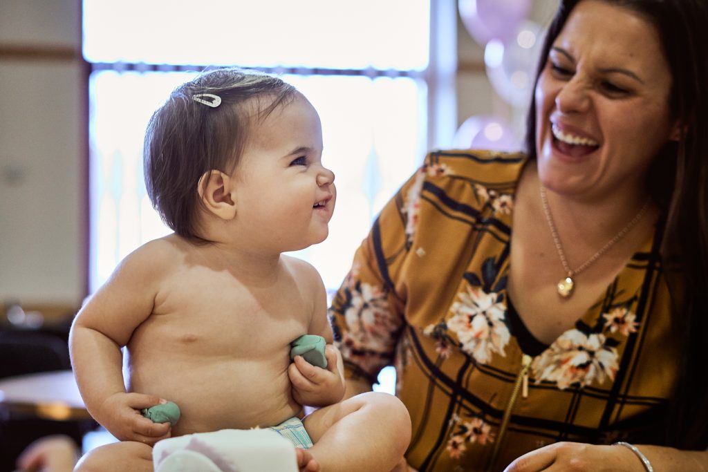 a cute baby girl sits in her cloth gnappies nappy holding bits of her christening cake and gives a cheeky smile to her mum who is laughing for their family photographer love gets sweeter