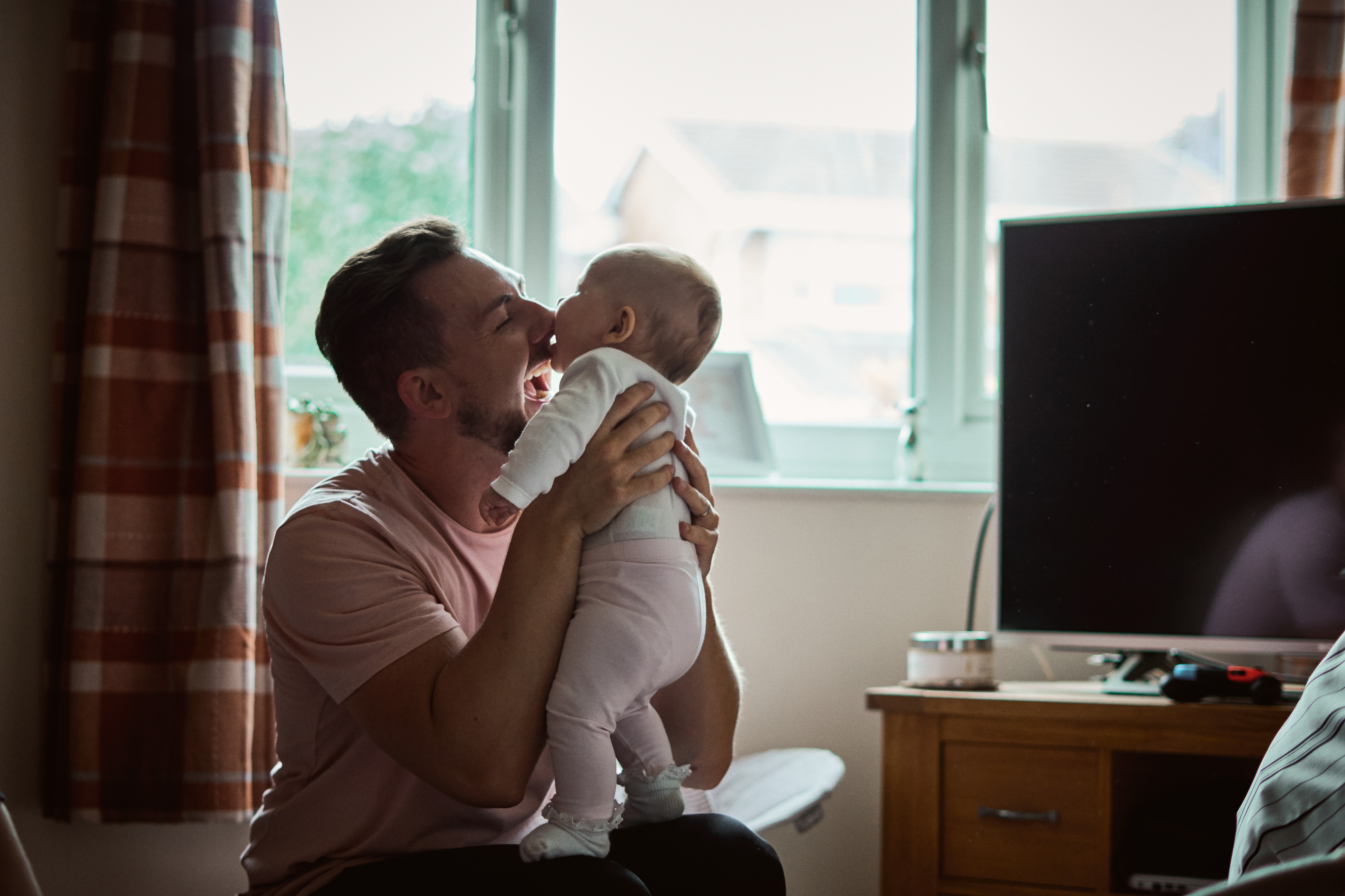 a dad crouches in front of a window and pulls his baby daughter in for a big kiss captured during a documentary lifestyle family shoot in their home in Lydiate