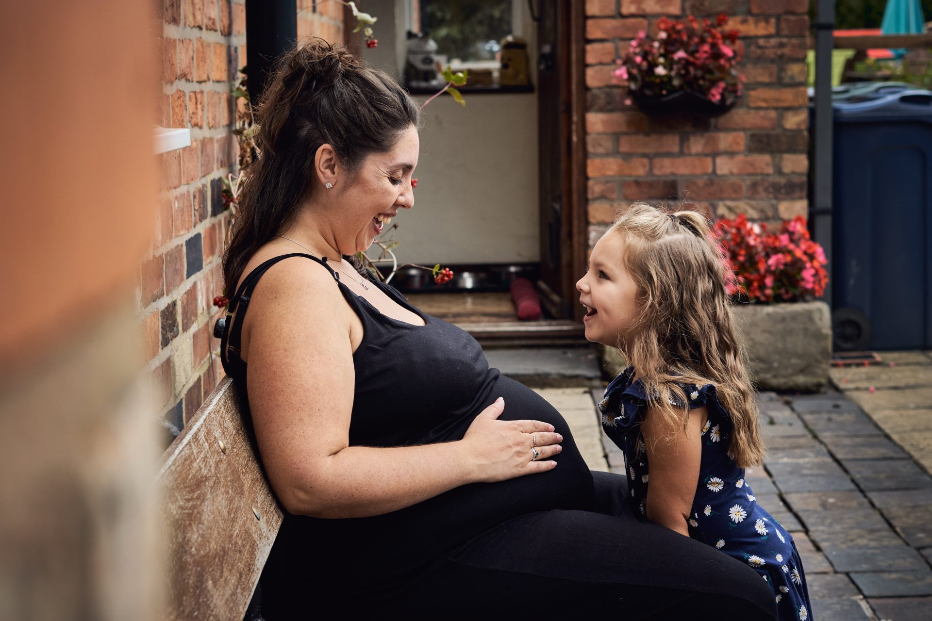 A mum sits on a wooden bench outside their family cottage in Newbrugh holding her baby bump and smiling down at her other daughter during a documentary style family maternity photoshoot by love gets sweeter family photography in Lancashire
