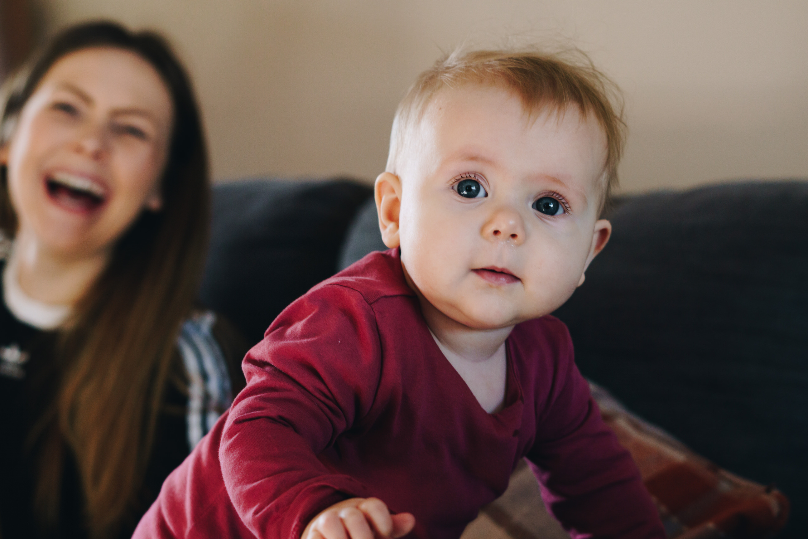 a baby girl leans towards the camera with her big blue eyes and her mum is seen laughing at her in the background during a home family photo shoot near Lancashire
