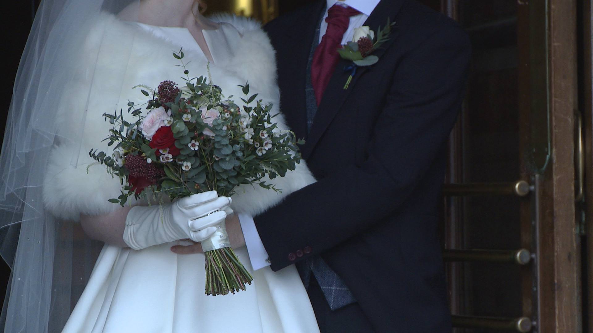 a close up of a white glove on the bride holding her rustic red and pink wedding bouquet as the groom puts his arms round her waist for their wedding photographer outside the villa at wrea green
