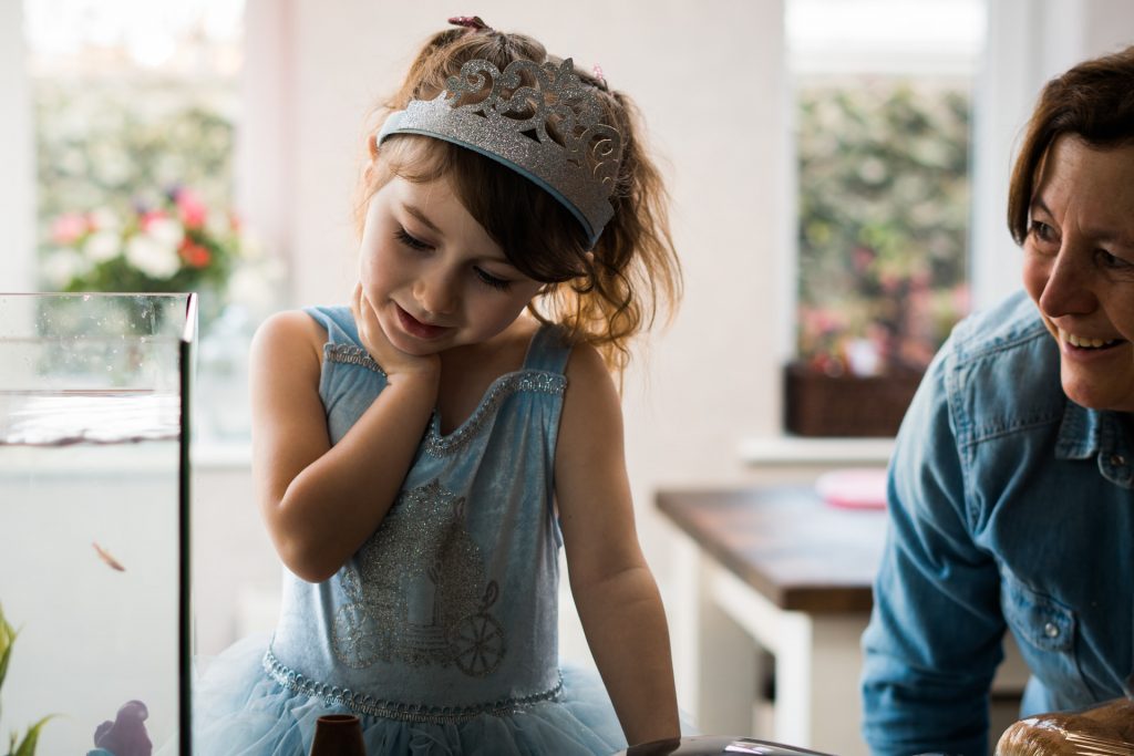 a little girl smiles with her nana at her new fish wearing a blue cinderella princess dress and silver crown ready for her birthday party at home in Burscough lancashire