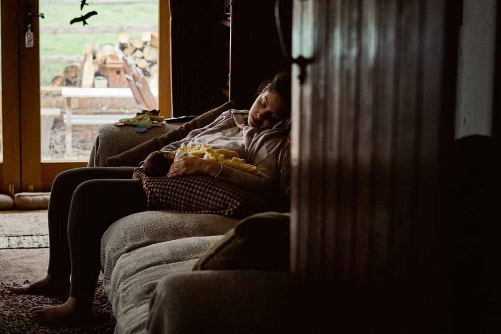 the photographer love gets sweeter peers through the doorway to capture a Mum and her newborn baby boy having a sneaky dose on the living room sofa after a feed during a home shoot in Newburgh Lancashire