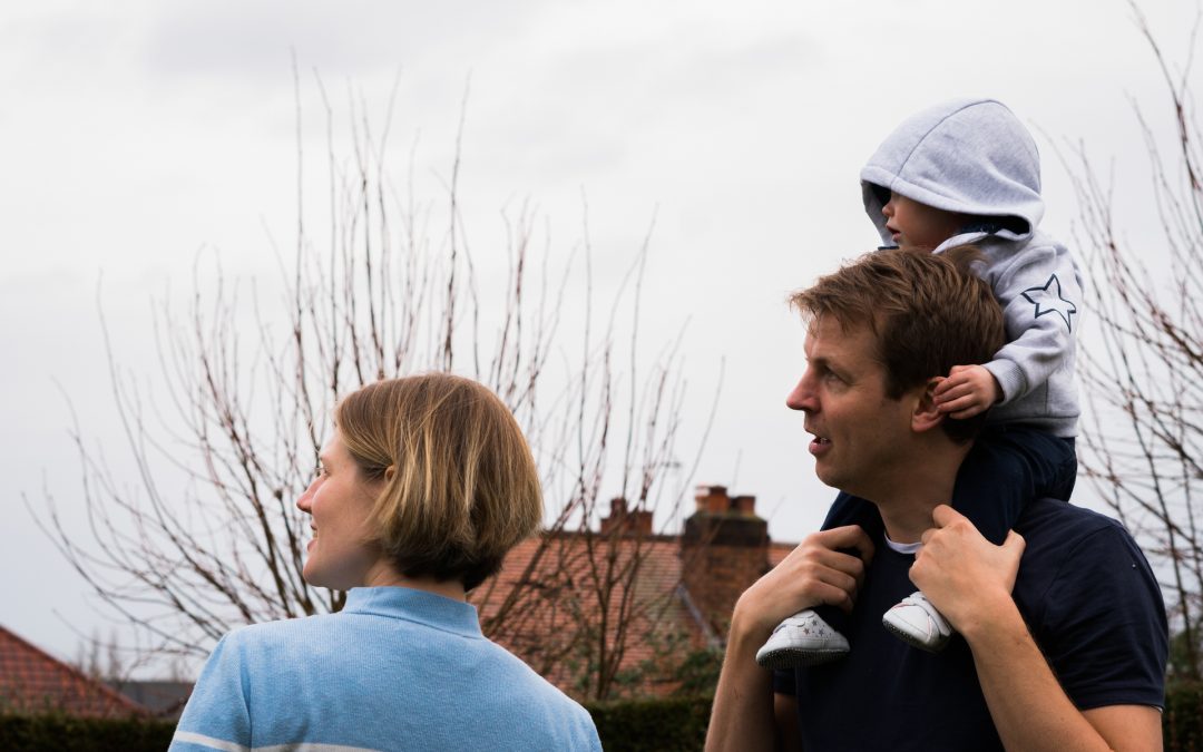 a little girl sits on her dads shoulders and holds on to his ears during a fun family shoot in their garden near Manchester