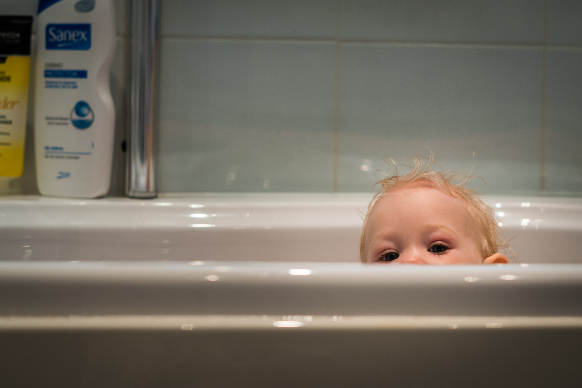 baby girl peers over the bath with her wet curly blonde hair during a relaxed documentary family photo shoot at their home in Hale near Manchester