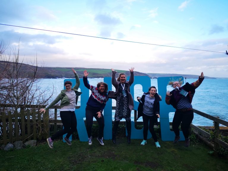 5 female wedding videographers jump in the air for a photograph in front of giant blue letters spelling RISE at a cove in Cornwall for the RISE wedding filmmakers retreat