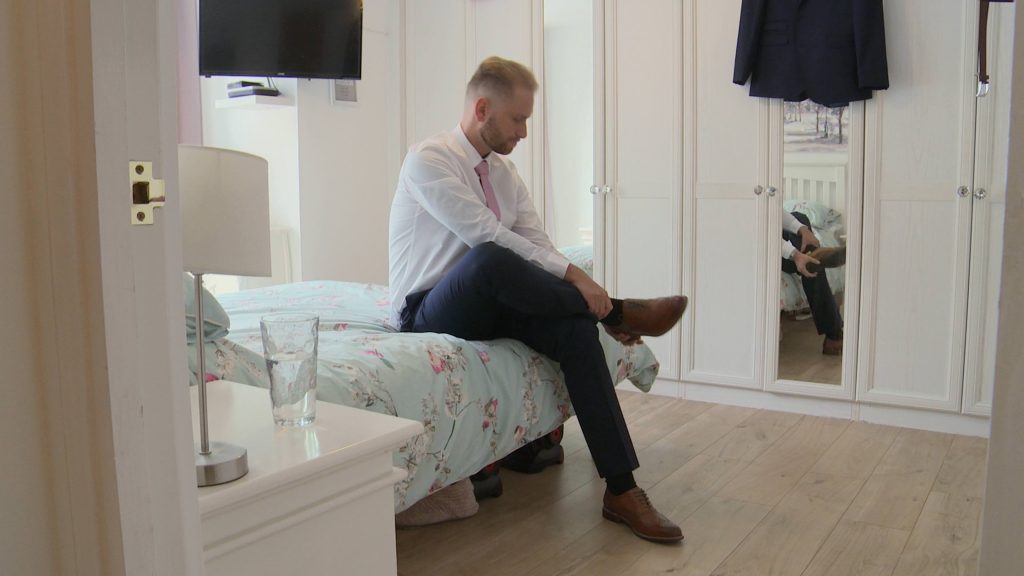 a still from the wedding video of a groom sitting on a bed at his home in Prenton tying his brown leather wedding shoes