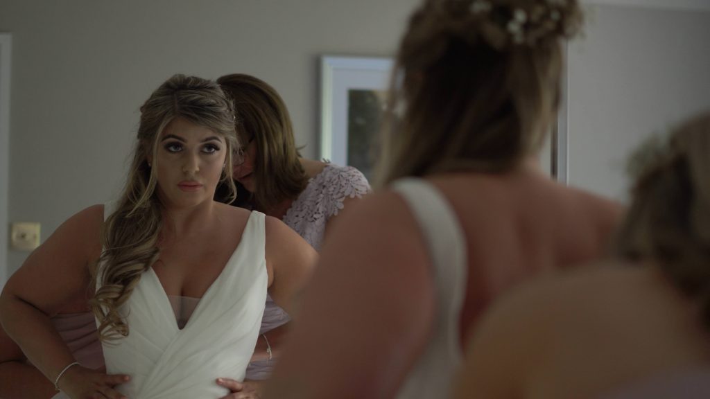 a still from the wedding video of a brides reflection in a wardrobe mirror getting her wedding dress fastened by her Mum in the wirral