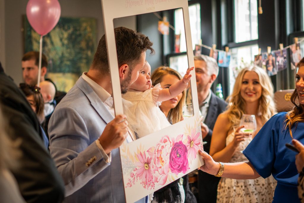 guests laugh as the little girl who got christened is held up to pose her a godparent inside a bespoke Instagram frame during her party at Site Pizzeria in Lancashire