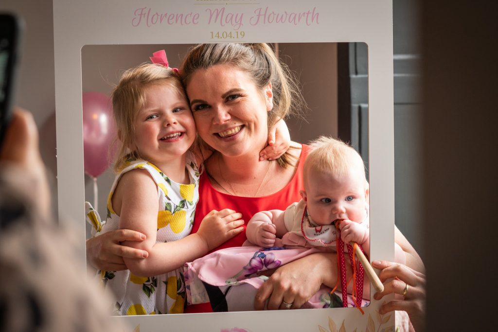 the family photographer takes a natural photo of a young family with two girls posing for someone holding a bespoke instagram frame for Florences christening