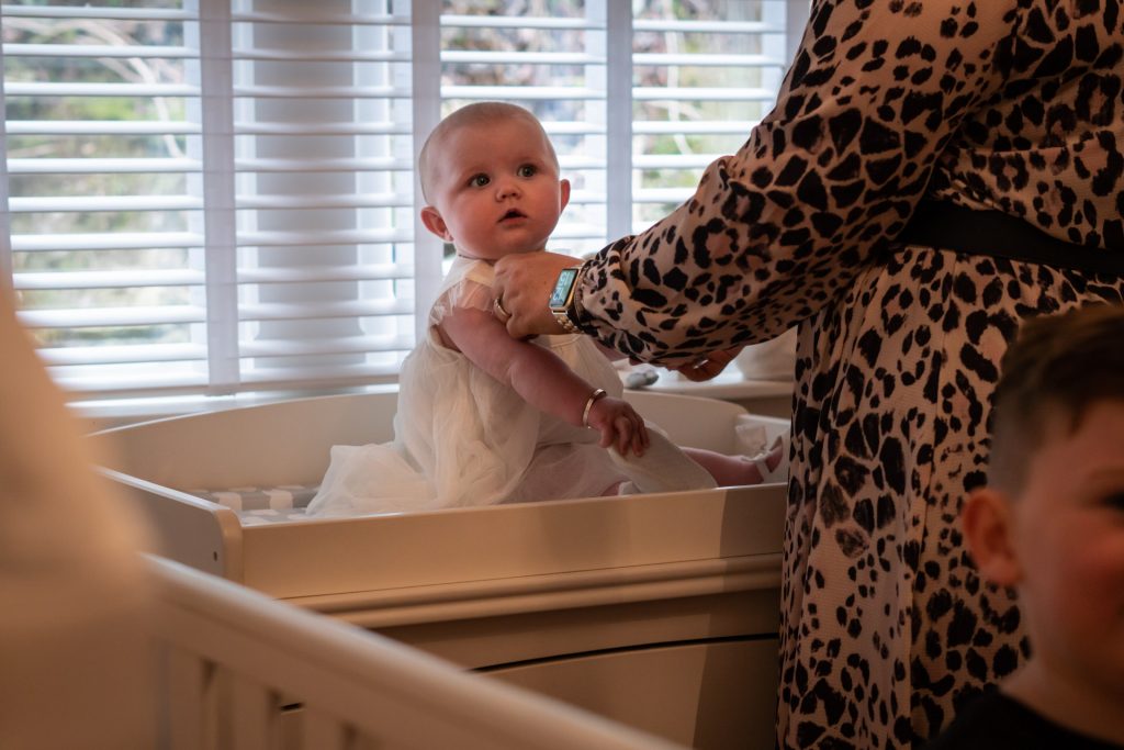 a little baby girl called Florence sits on her changing table in her nursery at home in Todmorden getting her white vintage style christening dress on as Love Gets Sweeter photographs the morning before her christening
