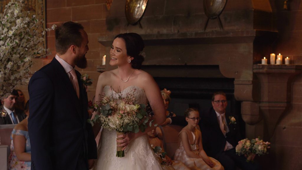 a bride and groom smile at each other during their peckforton castle wedding ceremony in front of the wedding videographer