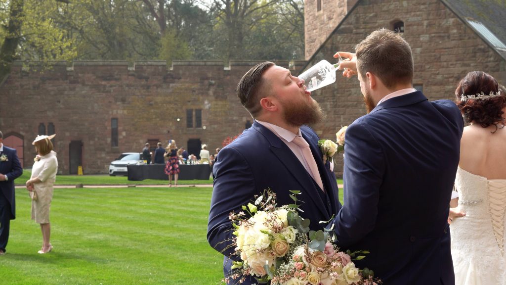 a groom helps his best man down the last of his beer during wedding photos on the lawn at Peckforton