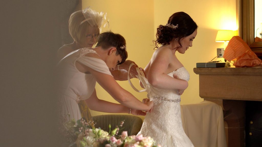 a still from the wedding video of a bride getting in to her wedding dress with the help of her Mum and sister in a bedroom suite at Peckforton Castle