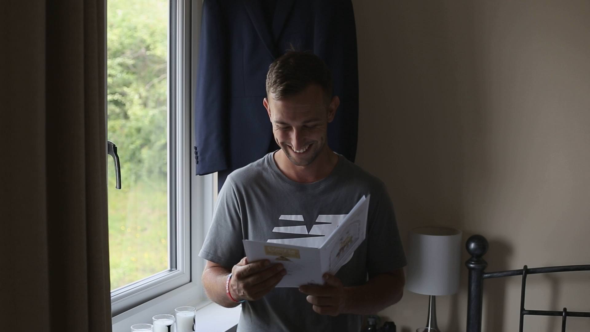 a groom stands by a window reading a card on the morning of his wedding