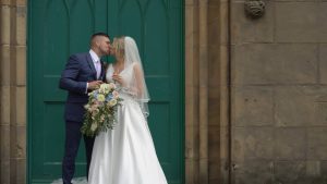 a full length wedding video still of a bride and groom kissing outside the green doors at St Johns Church