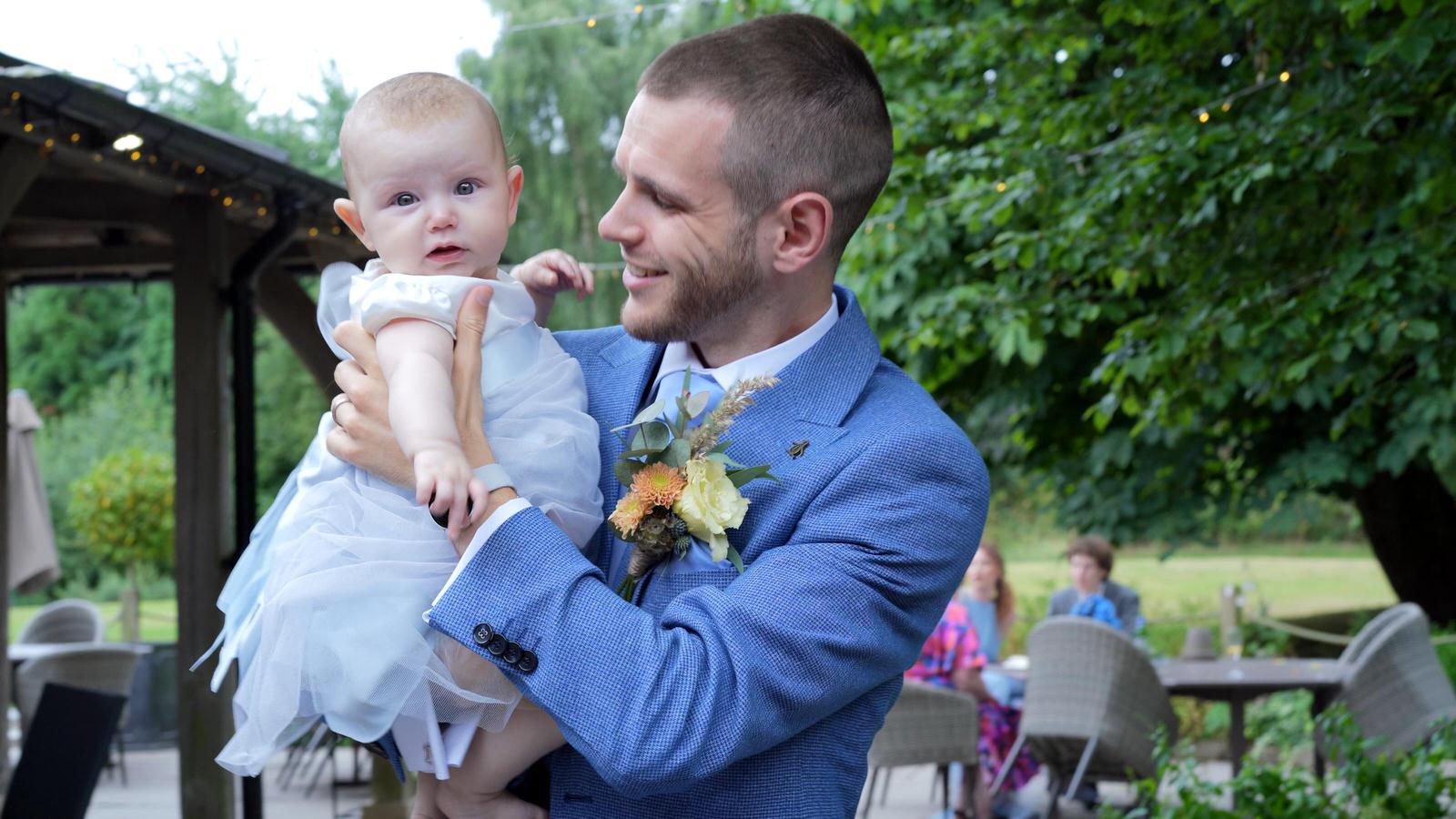 groom smiles at his baby daughter in flowergirl dress