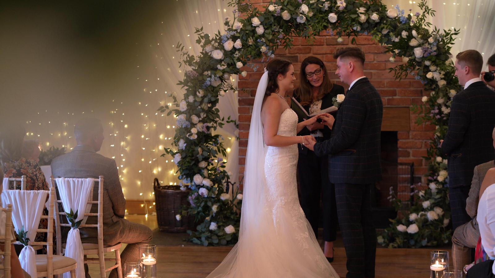 the couple exchange rings in front of a flower arch at Charnock Farm