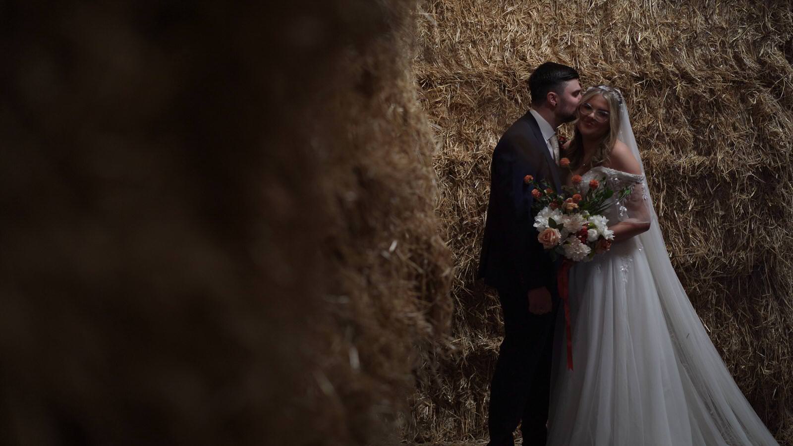couple stand in a pool of natural light by farm straw bales