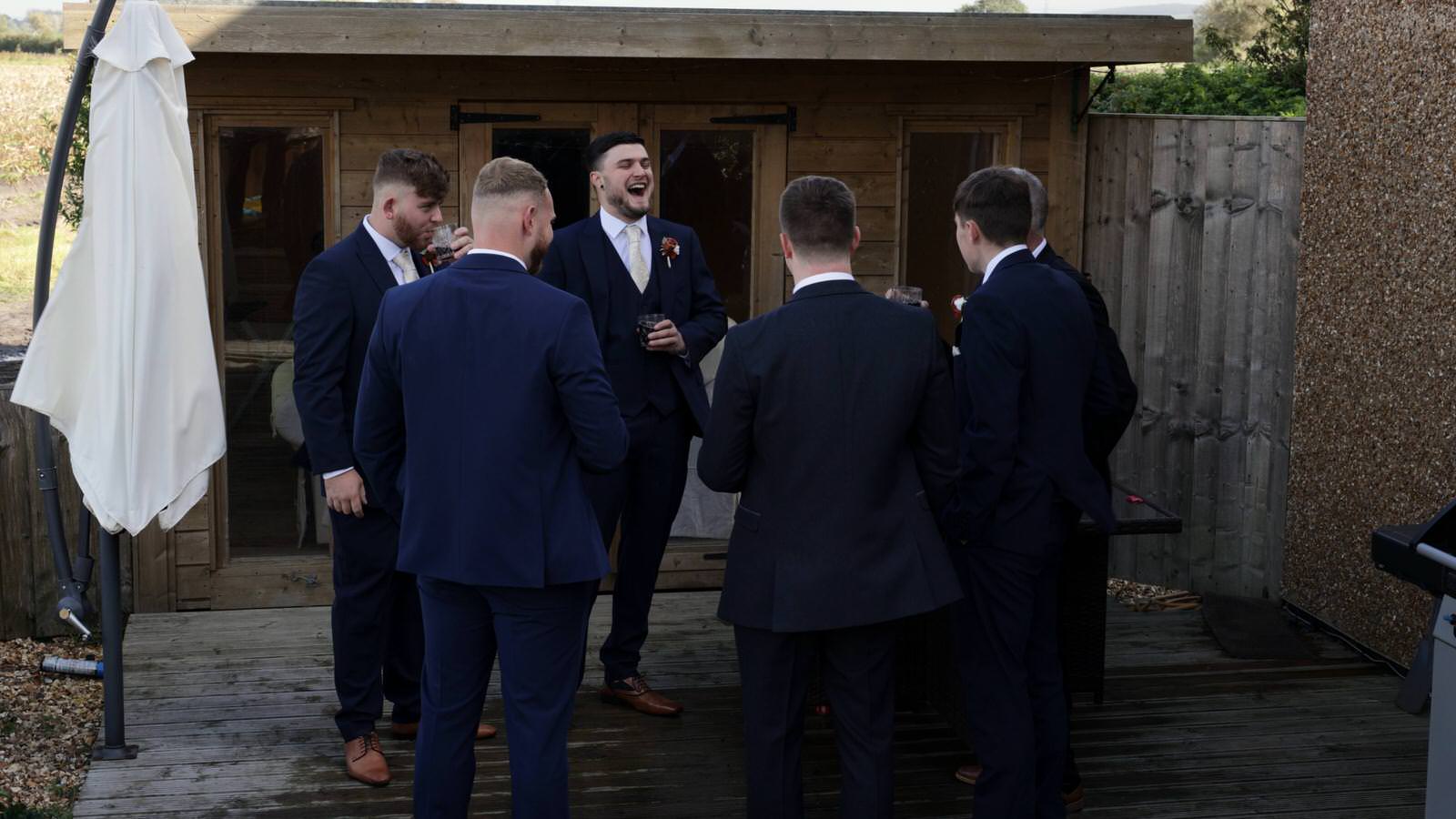 groomsmen stand and chat with beers outside farm house