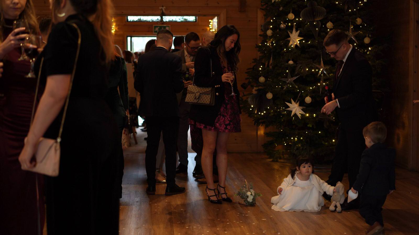 guests mingle by christmas tree and flower girl sits on floor