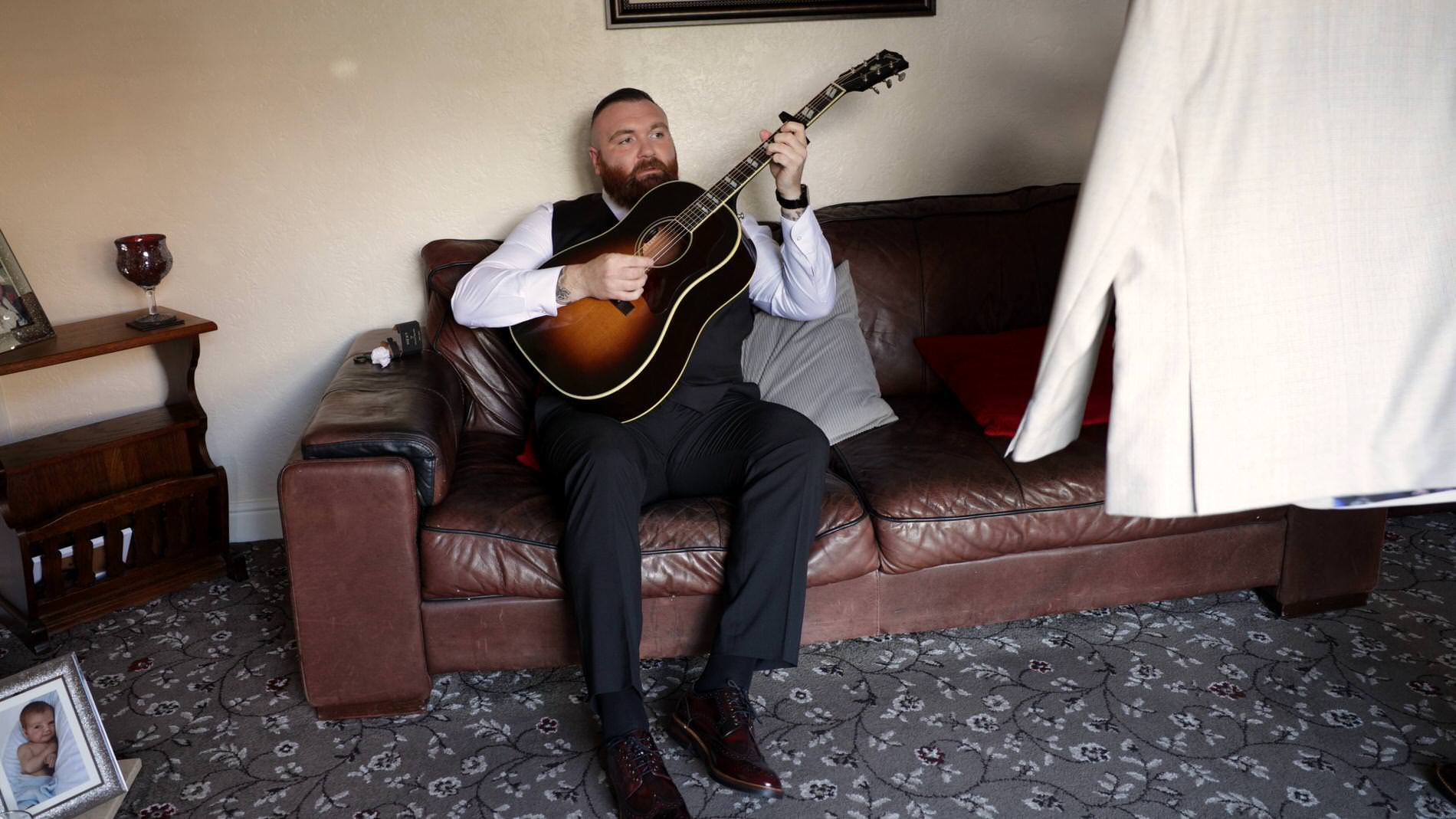 a groom sits casually on a sofa playing his guitar at home in Upholland
