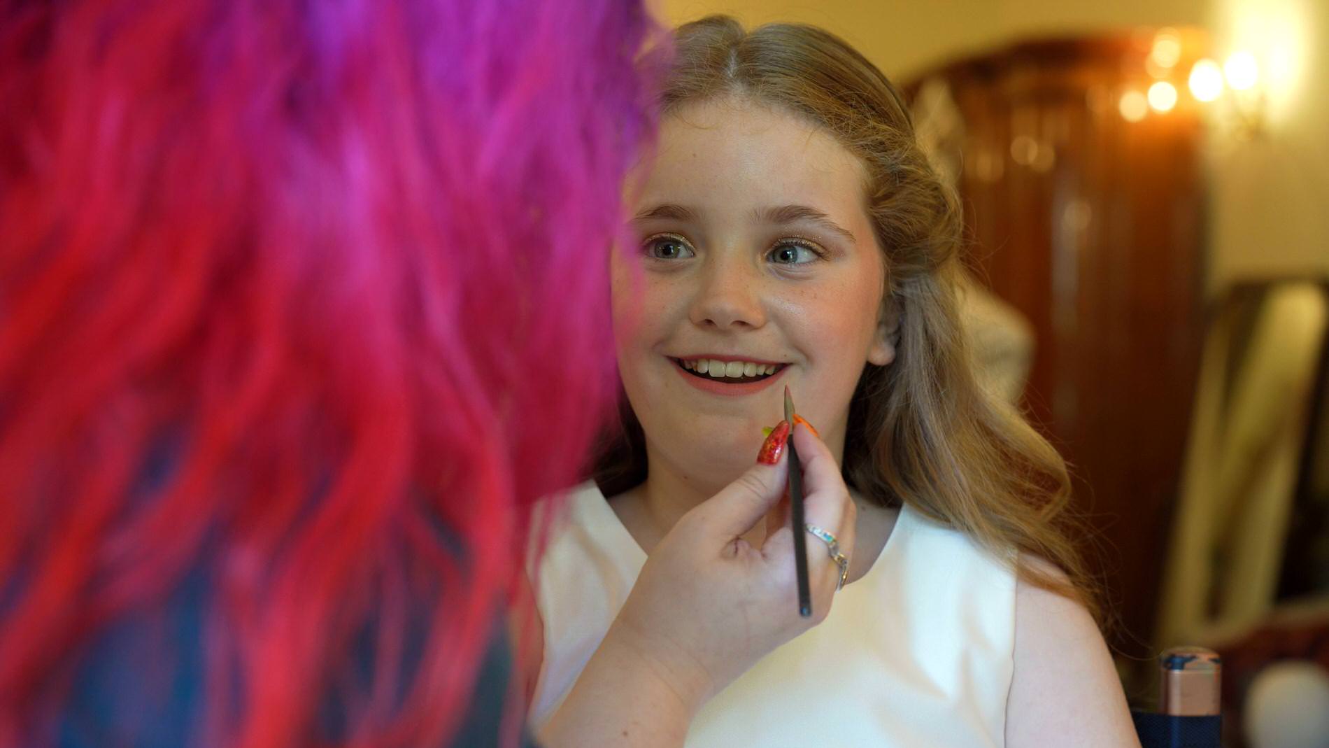 flowergirl smiles as she tries lipstick for the first time at a wedding