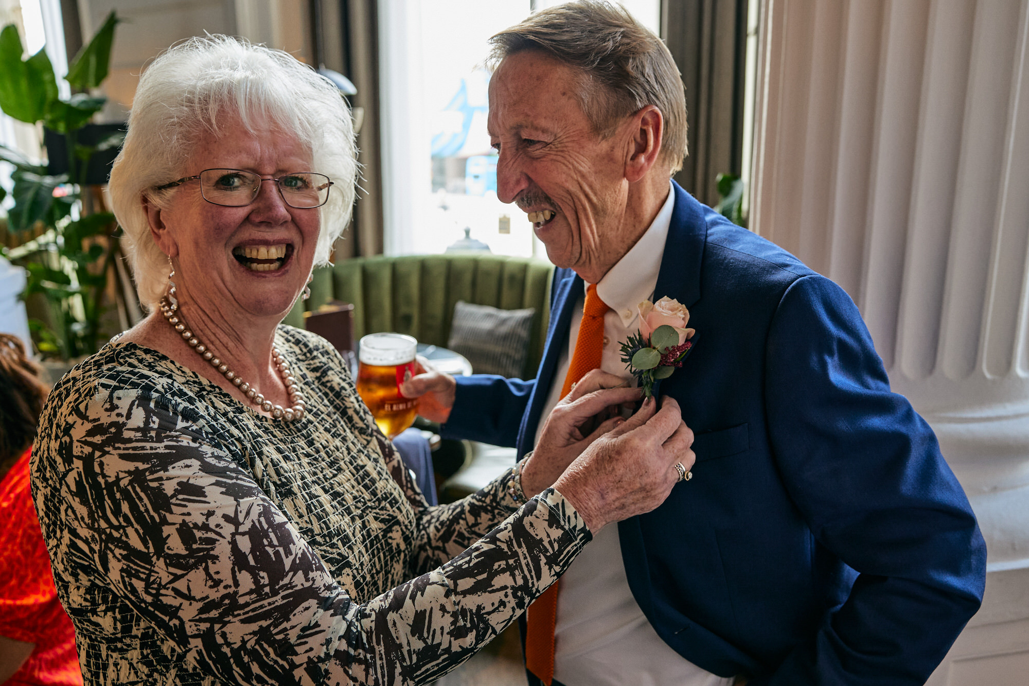 the grooms parents laugh at the camera as they try to do a buttonhole