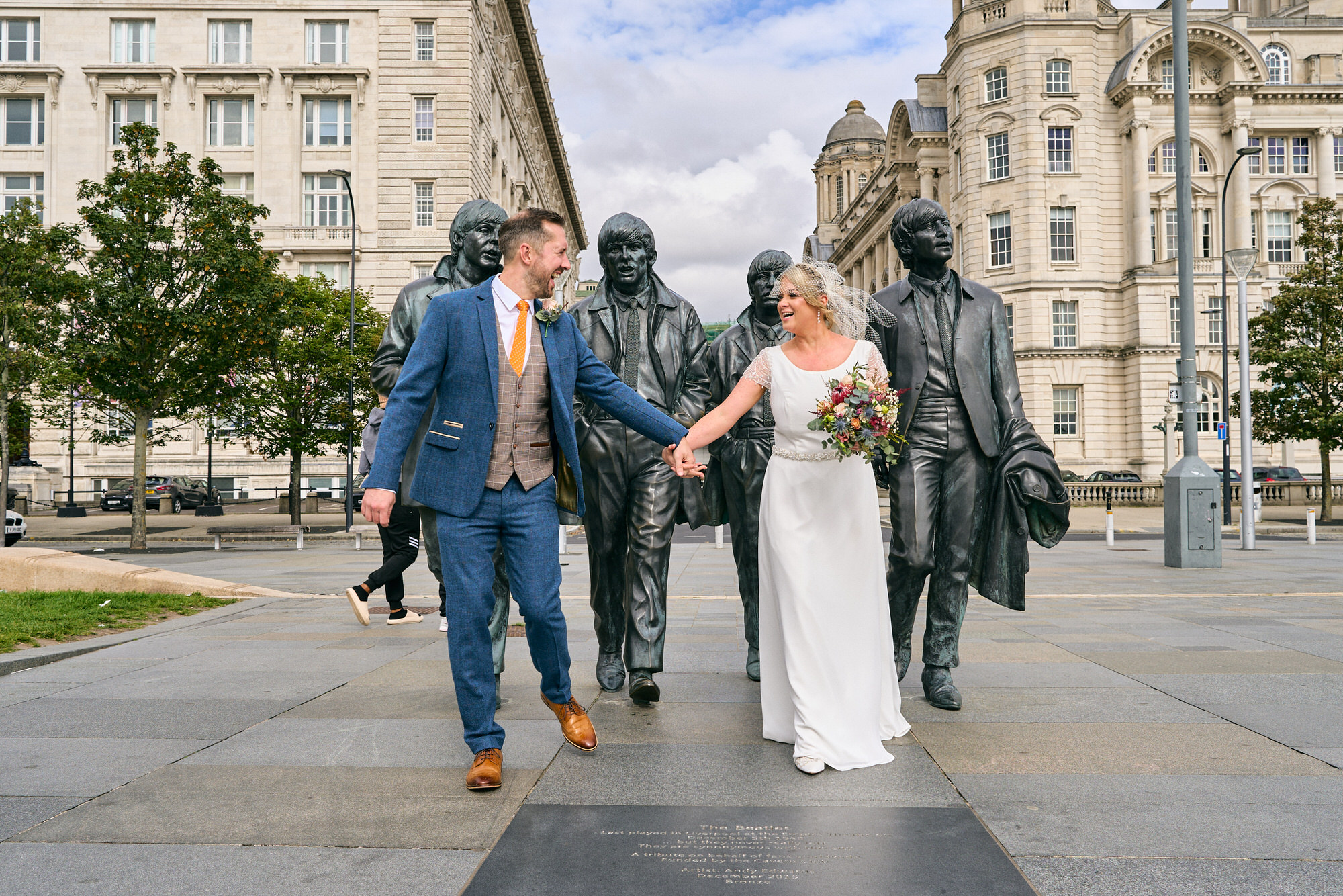 a couple walk and laugh towards the photographer with the Beatles Statue on the docks