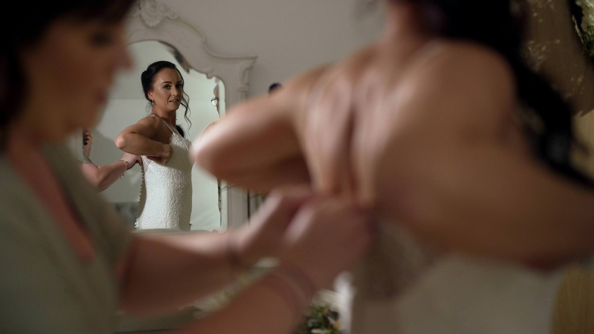 a bride glances at herself in the mirror as she gets her wedding dress on