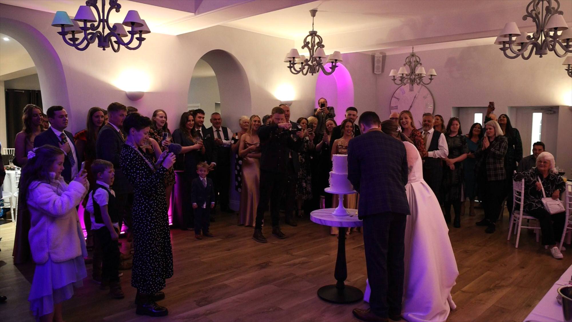 a wide video still of a couple cutting their wedding cake in front of guests