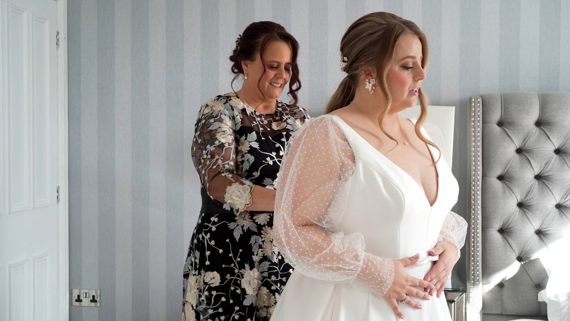 a bride has help from her mum putting on her wedding dress with spotty sheer sleeves