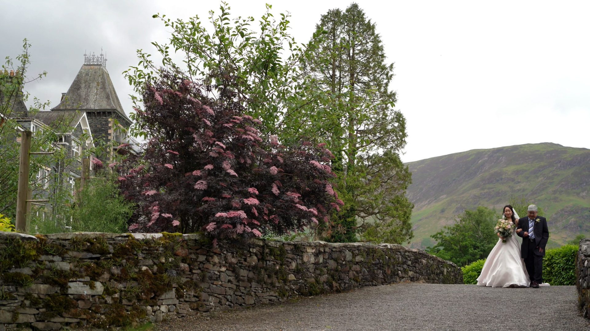 a wide shot of a bride walking to the outside ceremony with lodore falls hotel in the background