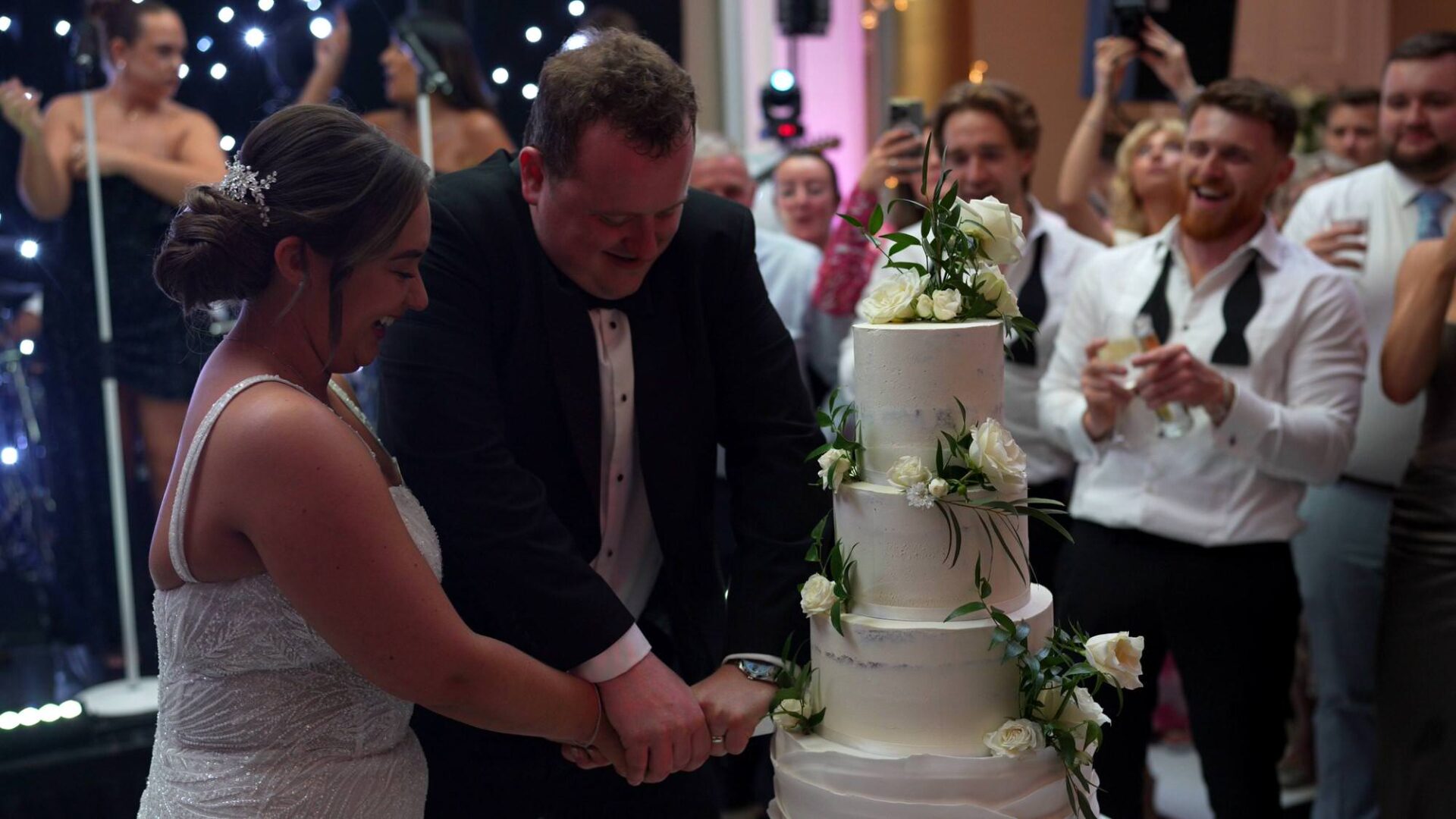 the groom needs to use a bit of force to cut the wedding cake as guests laugh