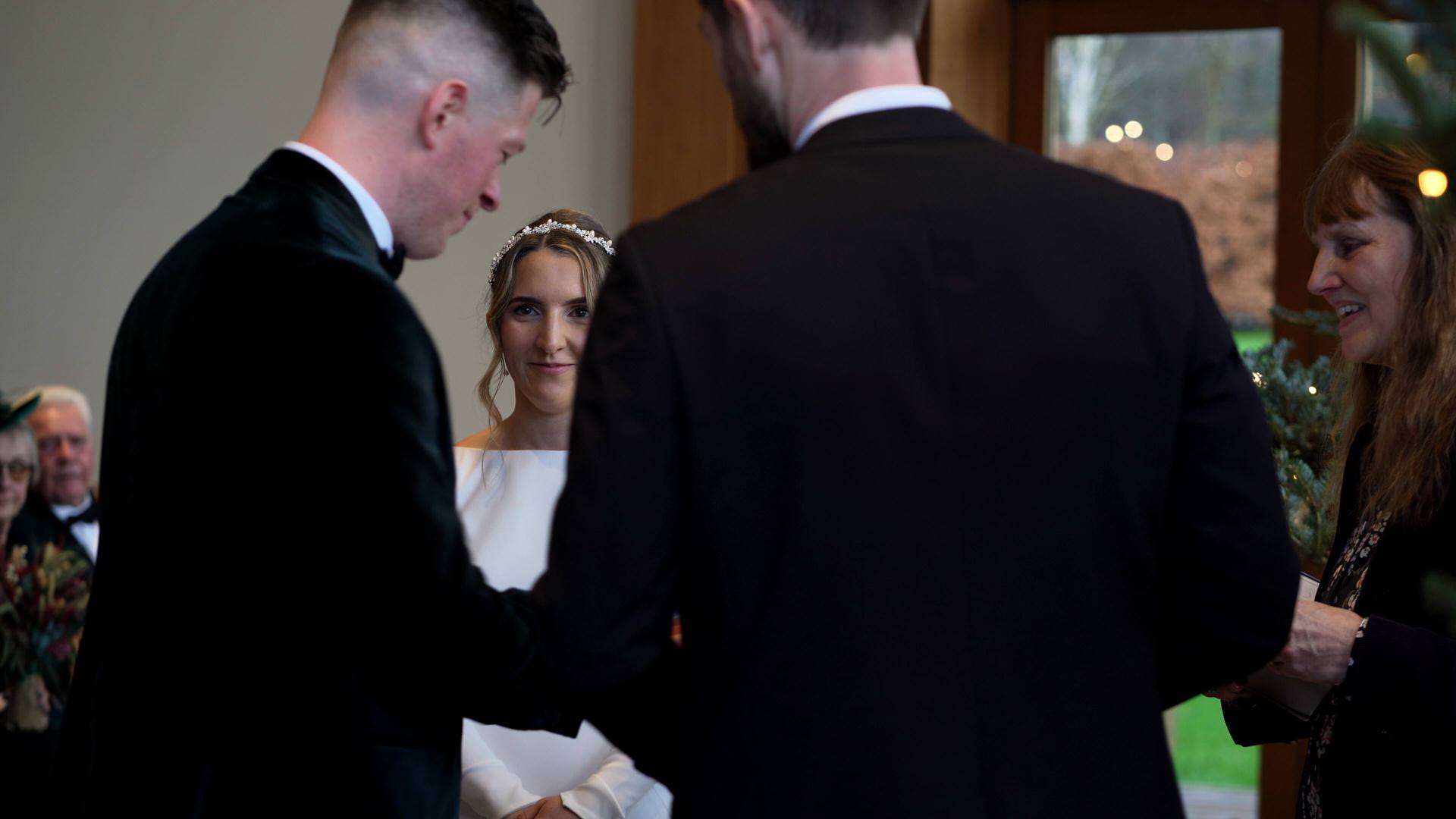 a natural moment of the bride smiling as the groom takes the wedding ring from the best man