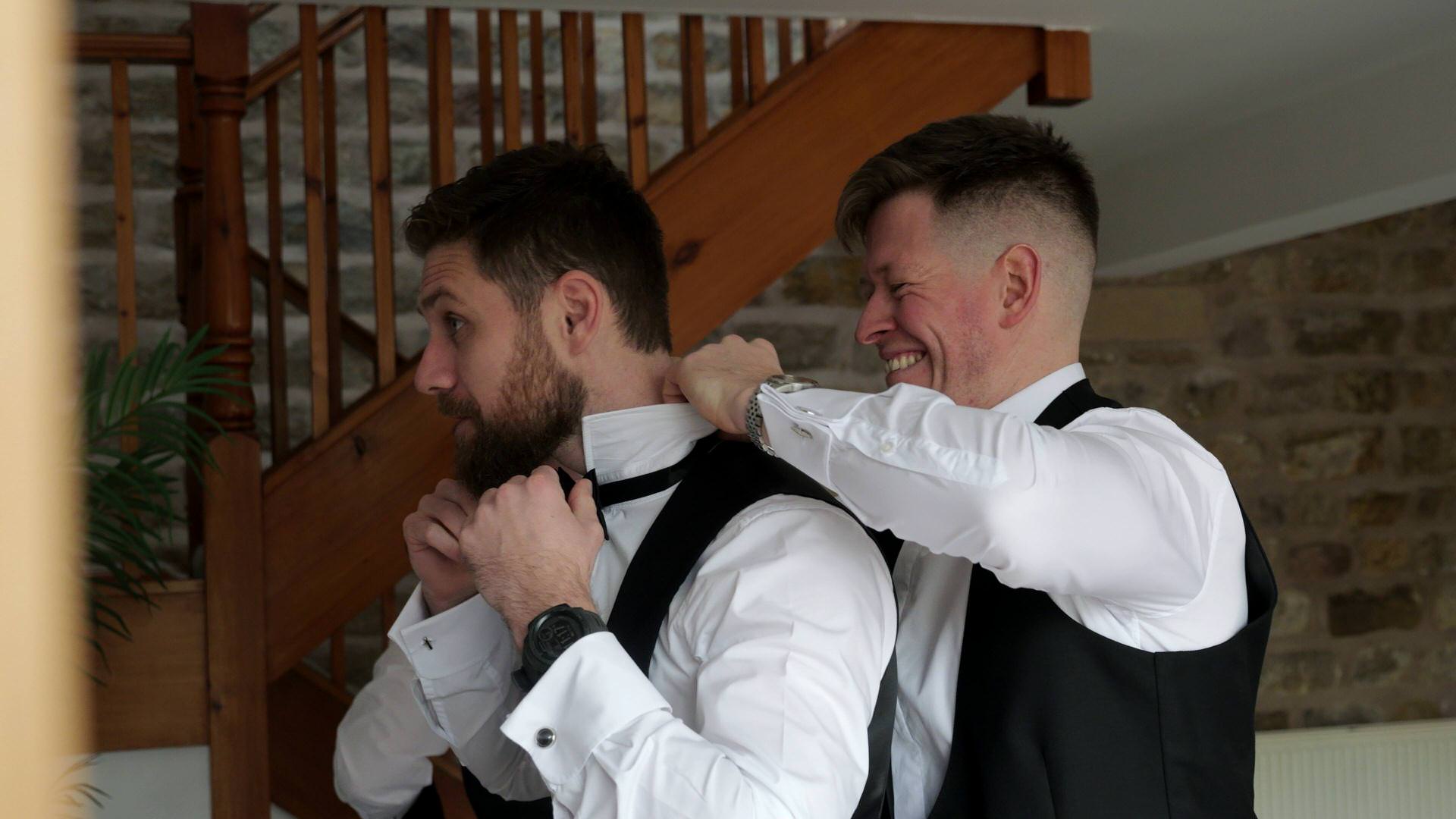 a groom laughs with his best man getting a black bow tie on