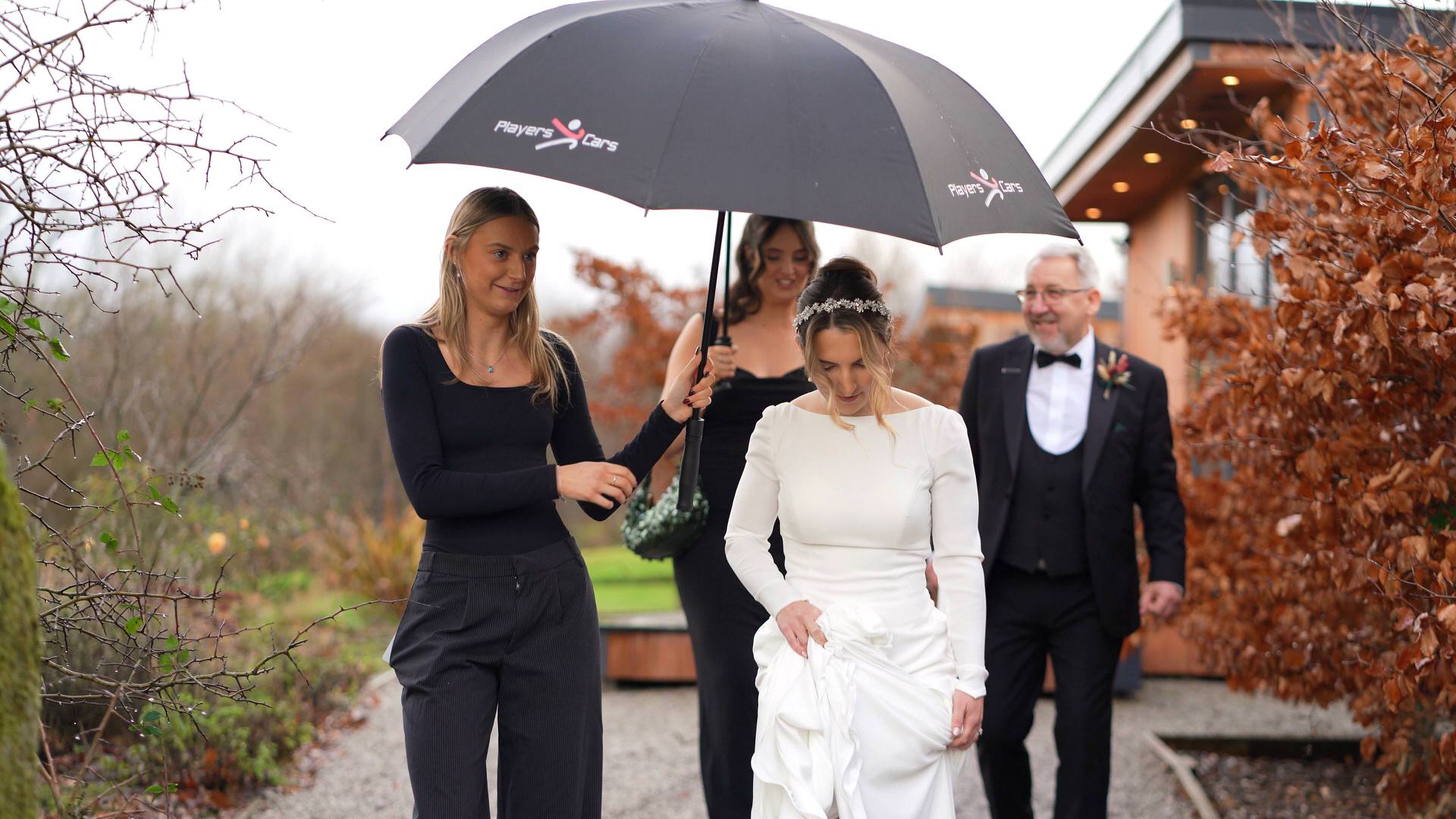 a videographer captures the bride leaving for the Out Barn wedding ceremony under an umbrella