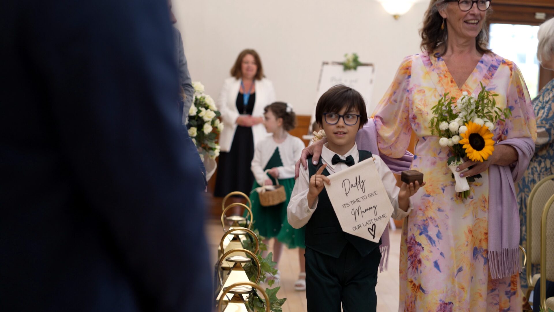 a page boy walks down the aisle with a cute handmade sign for his dad