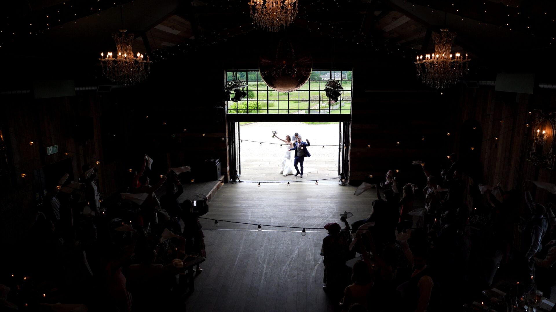 Candid documentary style wedding videography of a bride and groom entering the luxury rustic barn at Wharfedale Grange through the large glass doors as wedding guests wave napkins. Captured from high on the mezzanine