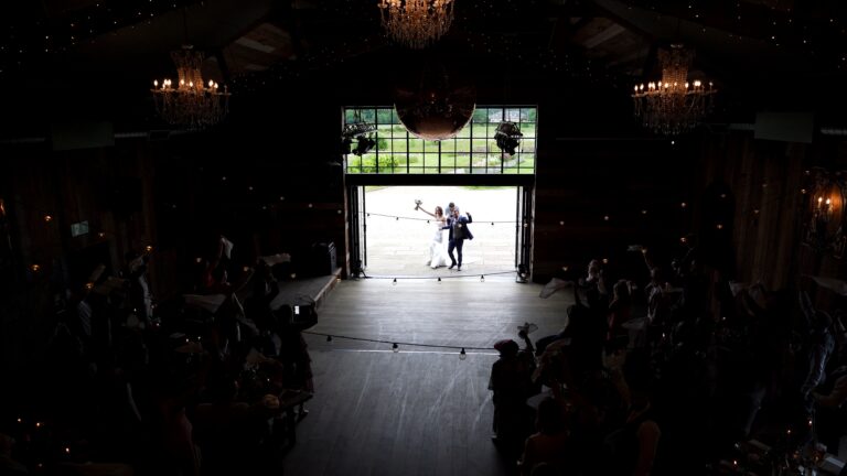 Candid documentary style wedding videography of a bride and groom entering the luxury rustic barn at Wharfedale Grange through the large glass doors as wedding guests wave napkins. Captured from high on the mezzanine