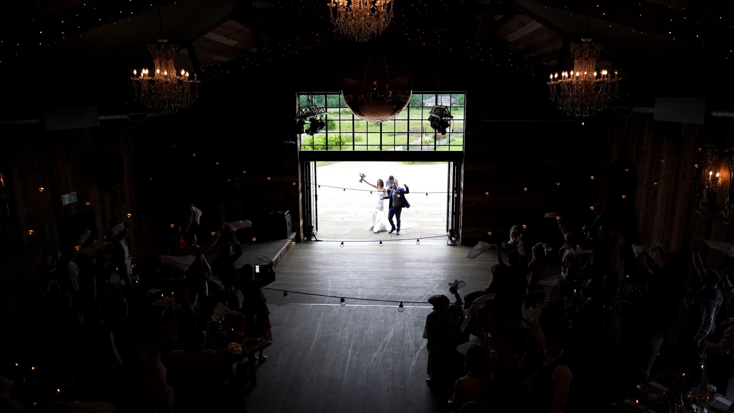 Candid documentary style wedding videography of a bride and groom entering the luxury rustic barn at Wharfedale Grange through the large glass doors as wedding guests wave napkins. Captured from high on the mezzanine
